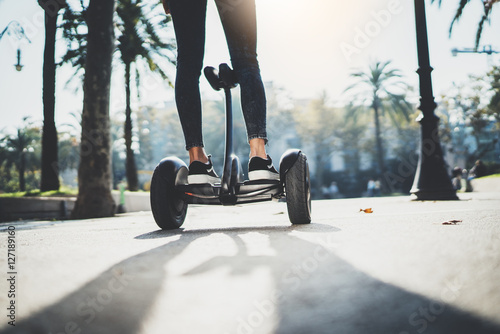 Back view of young hipster girl balancing on electric hoverboard at the sunny park, active woman using and driving on segway outdoor, film effects, flare light