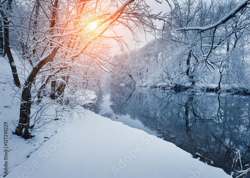 Winter forest on the river at sunset. Colorful landscape with snowy trees, frozen river with reflection in water. Seasonal. Winter trees, lake, sun and blue sky. Beautiful snowy winter in countryside