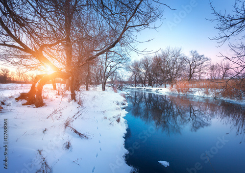 Winter landscape with trees, beautiful river at sunset. Winter forest in the evening. Season. Scenery with winter trees, water and blue sky. Christmas background with snowy forest. Reflection in water