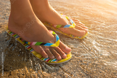 Female feet wearing flip flops. Water and sand under sunlight. One step away from sea. Visit a tropical island.