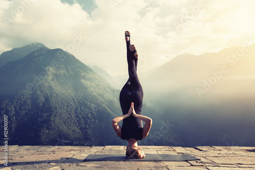Young woman doing complex Yoga exercise headstand with Namaste asana. Amazing Yoga landscape in beautiful mountains
