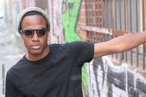 Fashion african man wearing a sunglasses, beanie, piercing and black tee over urban background in city alley 