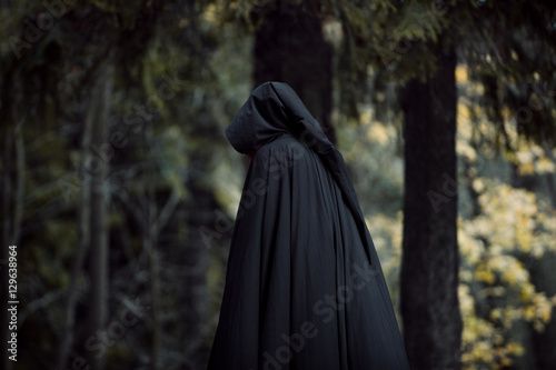 Young beautiful and mysterious woman in woods, in black cloak with hood, image of forest elf or witch