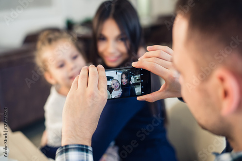 family, parenthood, technology, people concept - happy father taking photo of his little daughter and wife by smartphone having dinner at restaurant