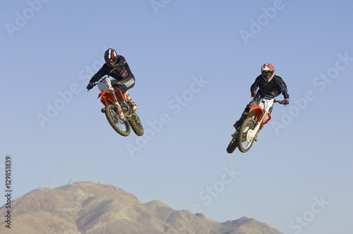 Two male motocross racers performing stunt in midair against clear blue sky