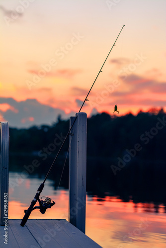 A fishing pole with frog bait rests on a dock at sunset over the lake.