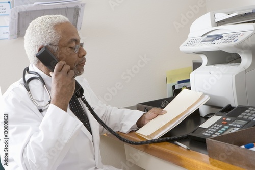 An African American senior male doctor holding document while using landline phone