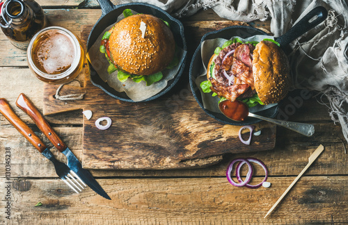 Homemade beef burgers with crispy bacon and vegetables in small pans and glass of wheat beer on rustic serving board over shabby wooden background, top view, copy space, horizontal composition