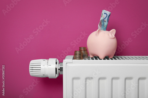 Savings concept. Piggy bank and money on heating radiator with temperature regulator on pink background