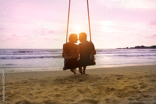 Romantic couple is sitting and kissing on sea beach on rope swing and looking at sunset horizon. Family vacation on honeymoon