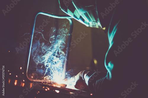 Industrial worker welding a loop made of round pipe on work table, producing smoke, sparks and reflections