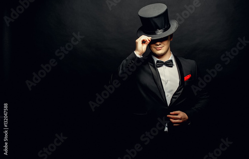 A magician in a black suit holding an empty top hat and magic wand isolated on black background