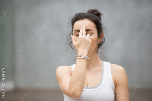Front view portrait of beautiful young woman wearing white tank top working out against grey wall, resting after doing yoga exercises, using nadi shodhana pranayama technique. Close up