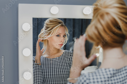 Woman in a beauty salon looks at her reflection in the mirror with lamps and checks hairstyle and makeup