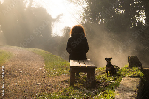 Donna seduta su una panchina con cane in una mattina con foschia lungo un sentiero