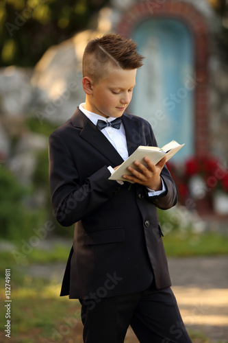 Śliczny model, chłopiec w garniturze z książeczką na tle kapliczki.