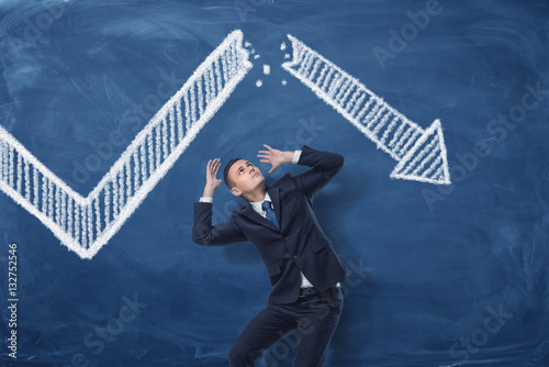 Businessman cowering on blue blackboard background with chalk drawing of white statistic arrow broken in half.