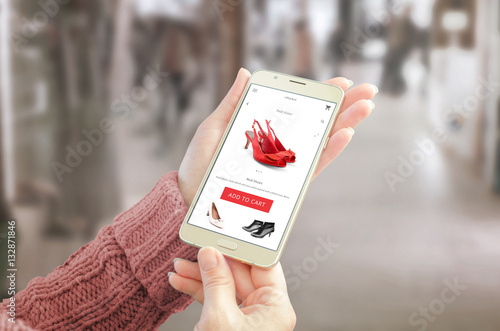 Woman holding smart phone with commerce web site. User friendly app interface with woman shoes. Shopping center in background.