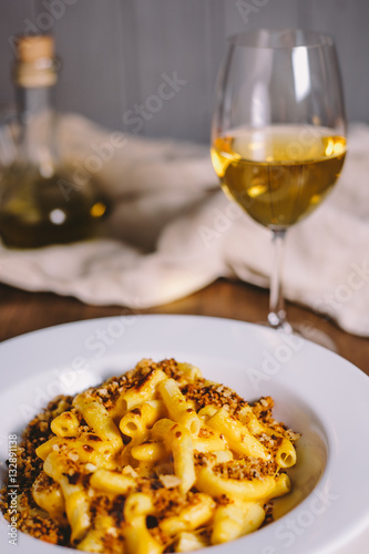 Cheese flavored rigatoni pasta in a white plate on a wooden table with kitchen appliances, glass of dry white wine and glass jug with olive oil on the old gray kitchen towel in the background