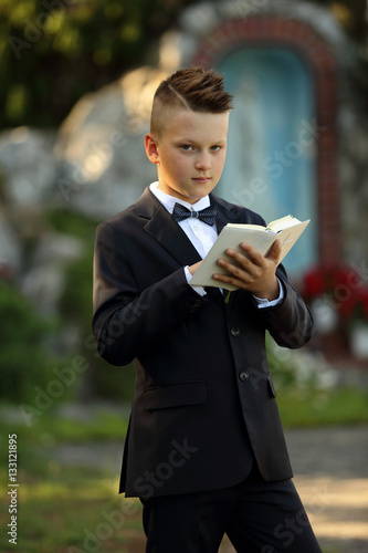 Pierwsza komunia, chłopiec w garniturze z książeczką na tle kapliczki.
