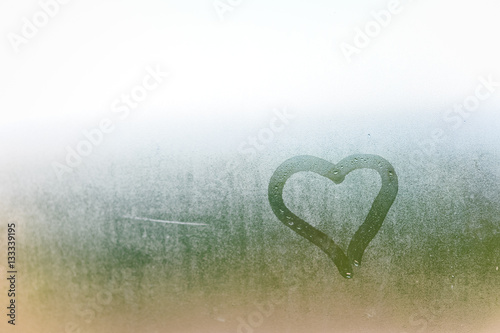 Abstract blurred love heart symbol drawn by hand on the wet frozen window glass with sunlight background.