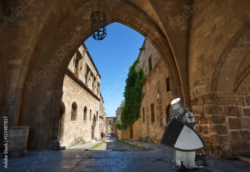 Medieval knight praying in entrance portal to the yard of Palace of the Grand Master of the Knights of Rhodes, Knights street lined with inns of different nationalities in the background
