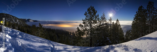Cold Winter Snowy landscape at night with cloud inversion covering city lights that glow underneath the cloud cover. Lit with moonlight and the sky has stars.