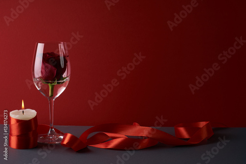 Wineglass with rosebud inside, candle and red ribbon on red background. Love card concept with copy space. Valentine's day theme