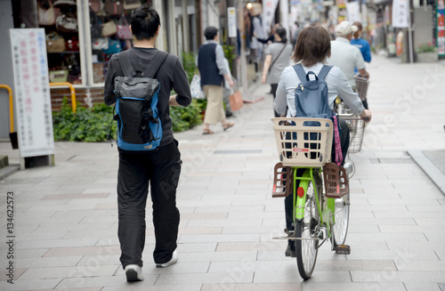 Idący pieszo młody mężczyzna z plecakiem oraz kobieta z plecakiem jadąca na rowerze z zamontowanym koszykiem do przewożenia dziecka