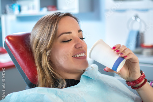 Smiling woman is taking a glass of water in dental office.