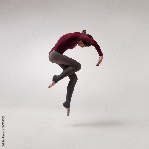 young beautiful woman dancer with long brown hair wearing maroon swimsuit jumping on a light grey studio background