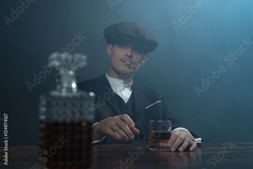 Retro 1920s english gangster sitting at table with whiskey. Peak