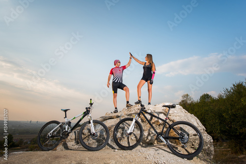 Young biker couple standing on a rock near bikes and giving high five against blue sky. Pink Kinesio tape glued on the girl's hand.
