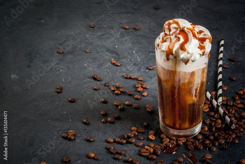 Cold coffee drink frappe (frappuccino), with whipped cream and caramel syrup, with straws and grains of coffee on a dark gray stone table, copy space
