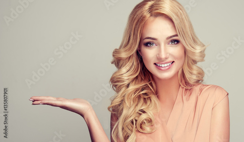 Blond kobieta z kręconymi włosami pokazuje swój produkt. .Piękna dziewczyna z pięknym uśmiechem skierowanym w bok. Prezentując swój produkt. Ekspresyjny wyraz twarzy