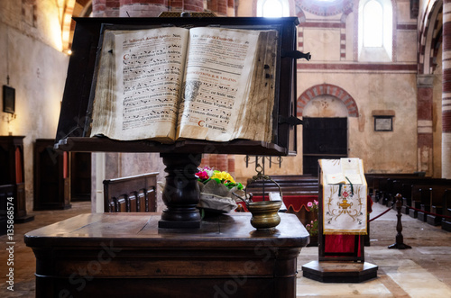 Ancient latin manuscript with religious chants and music sheets over a bookstand in the central nave of a romanesque church in an italian medieval abbey