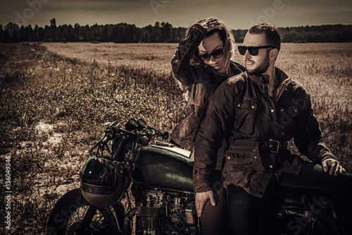 Young, stylish cafe racer couple on vintage custom motorcycles in field