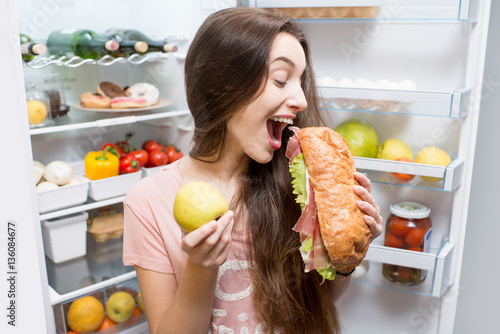Young woman choosing between apple and big sandwich standing in front of the refrigerator full of vegetables and fruits