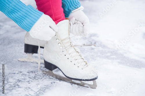 Woman tying white skates on the ice area in winter day. Weekends activities outdoor in cold weather.