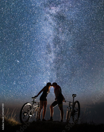 Romantic night. Young couple kissing under starry sky with mountain bicycles. Night landscape with colorful Milky Way. Bottom view. Copy space.