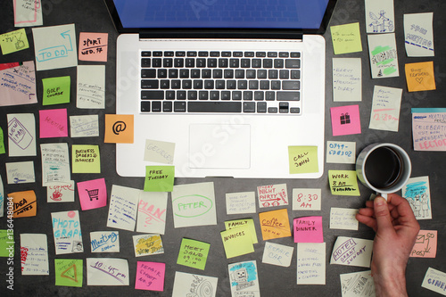 Top view on messy office desk with laptop, coffee and post it notes all around.