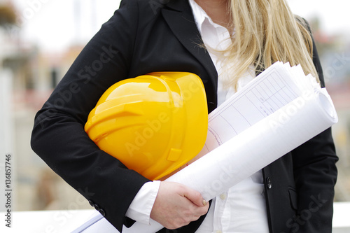 Junge Frau mit Helm und Plänen vor einer Baustelle