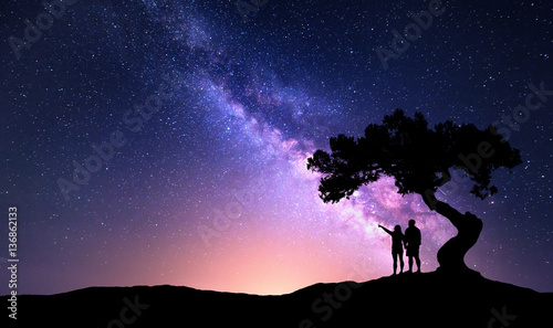 Milky Way with people under the tree on the hill. Landscape with night starry sky and silhouette of standing happy man and woman who pointing finger in starry sky. Milky way with travelers. Universe