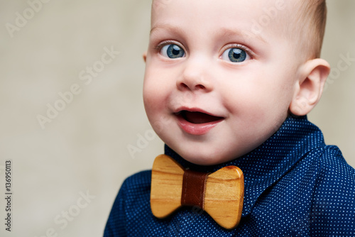 Cute caucasian baby boy wearing a fashionable blue shirt and a wooden bowtie all while looking straight into the camera with a positive smile on his face, along with his large blue eyes.
