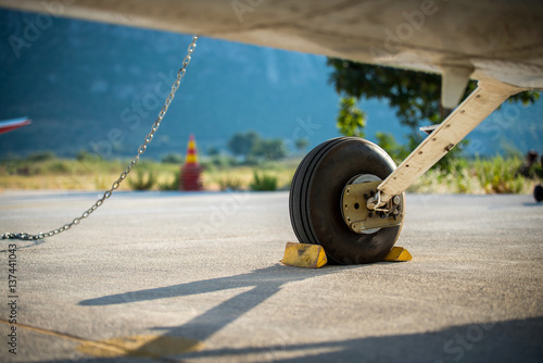 A rear landing gear and wheel chocks of a small aircraft on the ground with blurry nature and mountain in the background.