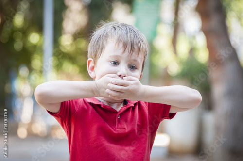 Serious little Caucasian boy closing his mouth with hands. Illustrative image for childhood trauma, child traumatic experience. Psychological assistance, children rescue. Silent cry for help. Stutter