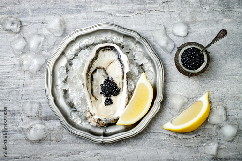 Opened oyster with black sturgeon caviar and lemon on ice in metal plate on grey concrete background. Top view, flat lay, copy space