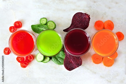 Four types of juice with scattered vegetables, above view over a white marble background