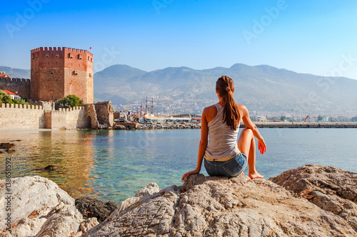 Young woman look at Kizil Kule tower in Alanya peninsula, Antalya district, Turkey, Asia. Famous tourist destination with high mountains. Part of ancient old Castle. Summer bright day