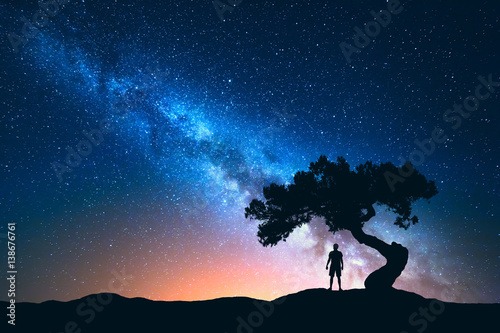 Night landscape with Milky Way and silhouette of man under the tree growing from the rock on the mountain. Nature background with starry sky and beautiful galaxy. Blue Milky Way and man. Universe